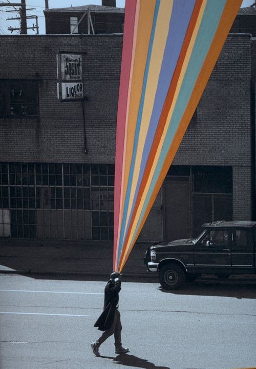 A man walking down a grey street carrying a huge rainbow-coloured banner