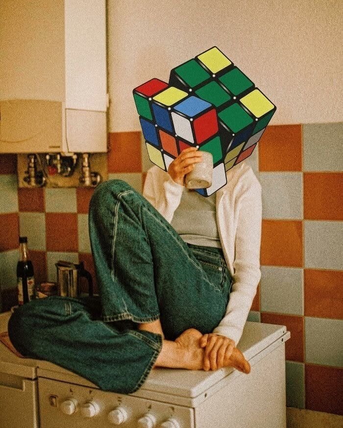 A woman with a Rubik's cube for a head, sitting barefoot on a kitchen stove