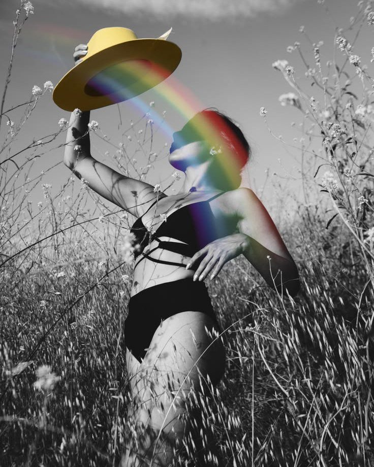 A woman in a field of grass raising a yellow hat, a rainbow of light falling across her